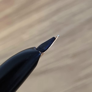 Może przedstawiać: Zbliżenie czarnej stal&oacute;wki pi&oacute;ra wiecznego ze złotym akcentem. Stal&oacute;wka jest pochylona, ukazując cienką końc&oacute;wkę i szczelinę, kt&oacute;ra umożliwia przepływ atramentu. Korpus pi&oacute;ra jest błyszczący i odbija światło.