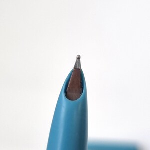 Puede incluir: Primer plano de la punta de una pluma estilogr&aacute;fica turquesa. El plum&iacute;n es plateado con una peque&ntilde;a bola en la punta. El cuerpo de la pluma es de color turquesa liso. El plum&iacute;n es marr&oacute;n y plateado.