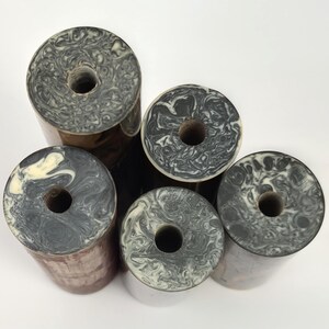 May include: Five cylindrical blanks arranged in a cluster, each featuring a distinct marbled pattern of black, grey, and white swirls. Every cylinder has a centred circular hole running through its length, showcasing a smooth, polished finish.