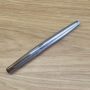 Puede incluir: Una taza de cer&aacute;mica blanca con un asa negra y el texto "But First, Coffee" en negrita negra. La taza est&aacute; sobre una superficie de madera. La taza mide aproximadamente 10 cm de alto.