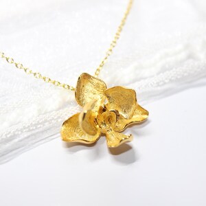 May include: A gold-coloured necklace with an orchid pendant. The pendant is a three-dimensional flower with textured petals, suspended from a delicate gold chain. The necklace is set against a white background.