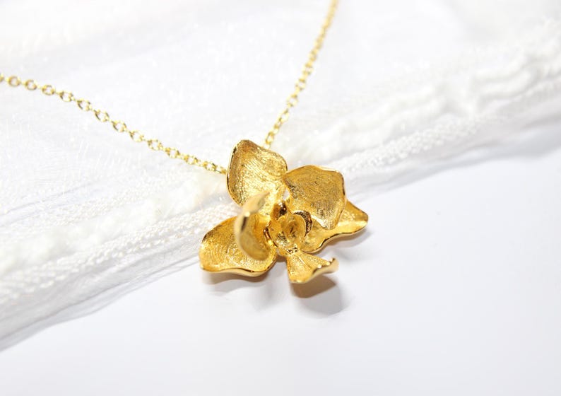 May include: A gold-coloured necklace with an orchid pendant. The pendant is a three-dimensional flower with textured petals, suspended from a delicate gold chain. The necklace is set against a white background.