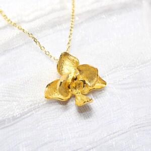 May include: A gold-coloured necklace with an orchid flower pendant. The pendant has textured petals and hangs from a delicate chain. The jewellery is displayed on a white, textured surface.