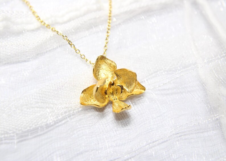 May include: A gold-coloured necklace with an orchid flower pendant. The pendant has textured petals and hangs from a delicate chain. The jewellery is displayed on a white, textured surface.