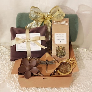 Könnte beinhalten: Ein Geschenkkorb mit einer grünen Decke, einem goldenen Band und einer braunen Box. Darin befinden sich ein dunkelviolettes Paket mit goldenem Band, ein Teebeutel, eine braune Blume, eine Libelle und ein goldumrandeter Gegenstand.