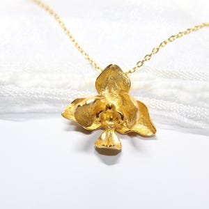 May include: A gold-coloured orchid pendant necklace. The pendant is a detailed, three-dimensional flower with textured petals. The necklace chain is delicate and also gold-toned. The background is a soft, white fabric.