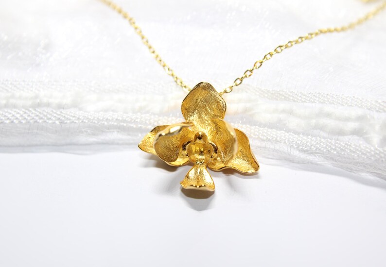May include: A gold-coloured orchid pendant necklace. The pendant is a detailed, three-dimensional flower with textured petals. The necklace chain is delicate and also gold-toned. The background is a soft, white fabric.