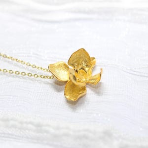 May include: A gold-coloured necklace with an orchid flower pendant. The pendant has textured petals and hangs from a delicate chain. The necklace is set against a white, textured background.