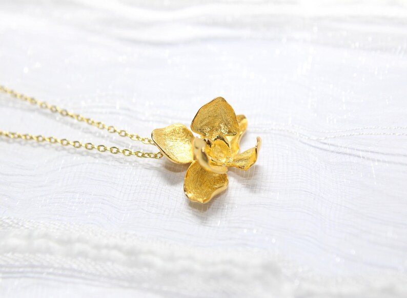 May include: A gold-coloured necklace with an orchid flower pendant. The pendant has textured petals and hangs from a delicate chain. The necklace is set against a white, textured background.