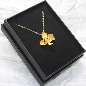 May include: A gold-coloured pendant necklace with a flower design, displayed in a black jewellery box. The necklace has a delicate chain, and the flower has detailed petals. The box is set against a white, textured background.