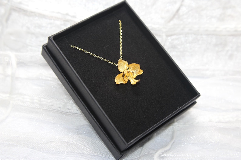May include: A gold-coloured pendant necklace with a flower design, displayed in a black jewellery box. The necklace has a delicate chain, and the flower has detailed petals. The box is set against a white, textured background.