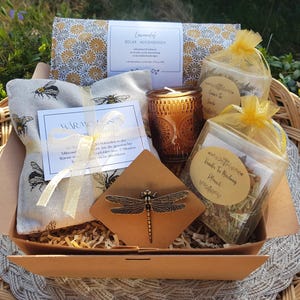 Puede incluir: Una cesta de regalo de relajación con una almohadilla térmica con estampado de abeja, una vela, bolsitas de té y una libélula decorativa. La cesta está forrada con papel y contiene una caja con estampado floral.