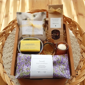 Könnte beinhalten: Eine Geschenkbox mit einem Augenkissen mit Bienenmuster, einer Packung Früchtetee Mischung Himbeere, einem gelben Lemongrass-Seifenstück, einer kleinen goldenen Dose, einer weißen Kerze in einem braunen Glas und einem Augenkissen mit Lavendelmuster.