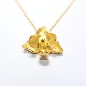 May include: A gold-coloured pendant necklace with a detailed orchid flower design. The pendant hangs from a delicate gold chain. The necklace is set against a white background, highlighting the jewellery.