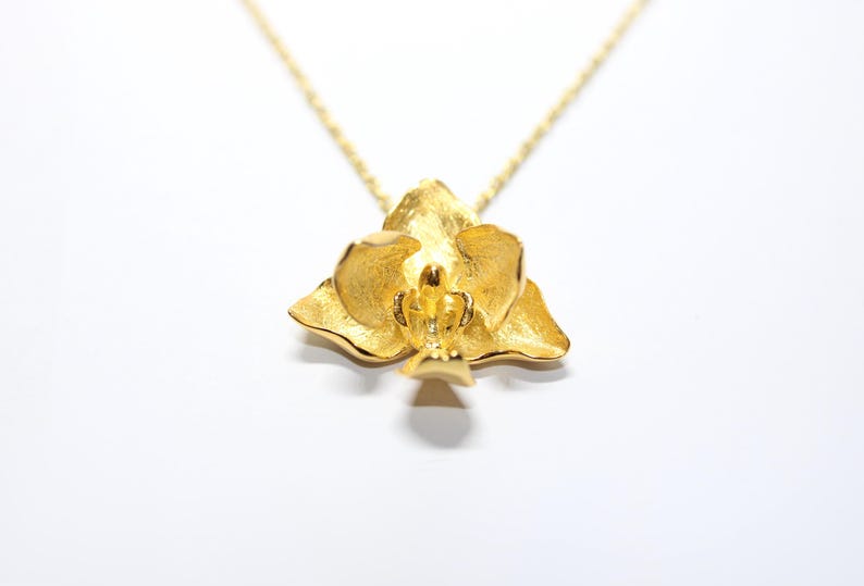 May include: A gold-coloured pendant necklace with a detailed orchid flower design. The pendant hangs from a delicate gold chain. The necklace is set against a white background, highlighting the jewellery.