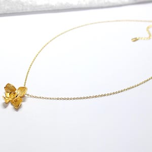 May include: A gold-coloured necklace with a floral pendant. The pendant is a detailed, three-dimensional flower design. The chain is a fine, adjustable link chain. The necklace is displayed on a white surface.