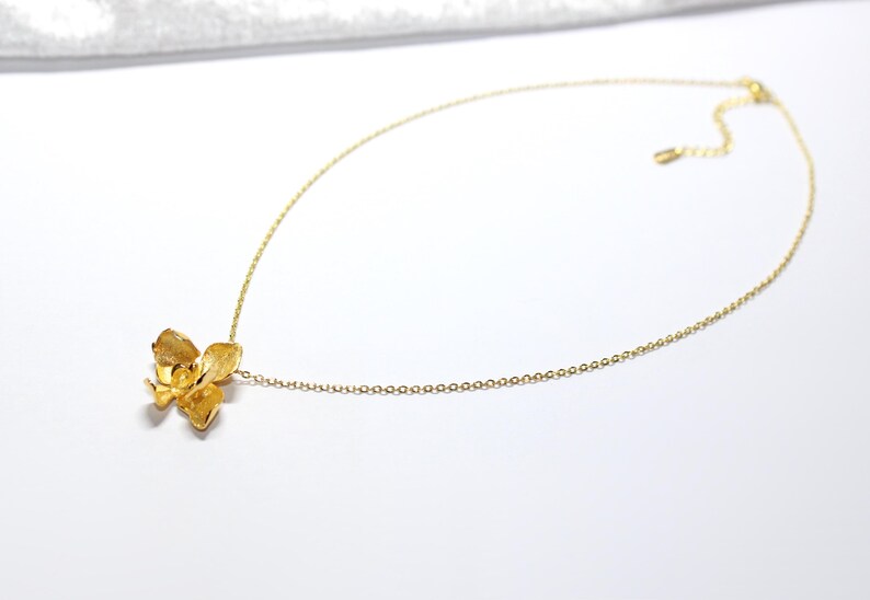 May include: A gold-coloured necklace with a floral pendant. The pendant is a detailed, three-dimensional flower design. The chain is a fine, adjustable link chain. The necklace is displayed on a white surface.