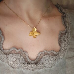 May include: A gold-coloured pendant necklace with a bird design. The bird pendant hangs from a delicate gold chain. The necklace is worn against a neutral background.