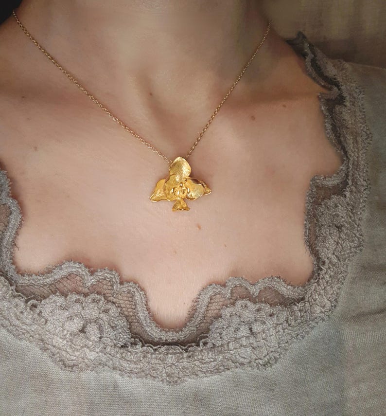 May include: A gold-coloured pendant necklace with a bird design. The bird pendant hangs from a delicate gold chain. The necklace is worn against a neutral background.