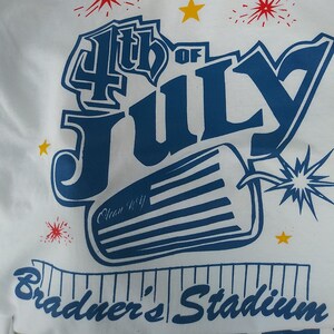 May include: White t-shirt with a blue and red graphic design that says "4th of July" with a firework design and the text "Bradmer's Stadium".