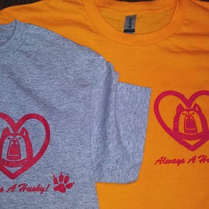 May include: Two t-shirts, one gray and one orange, with a red heart design featuring a husky dog and the text "Always A Husky!" printed on each.