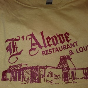 May include: A yellow t-shirt with a maroon print of a building and the text "L'Aleoive Restaurant & Lounge".