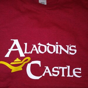 May include: Red t-shirt with white text that reads "Aladdin's Castle" and a yellow cartoon genie lamp.