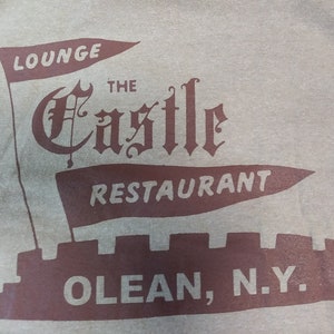 May include: A light gray t-shirt featuring a maroon graphic of a castle with flags that read "LOUNGE" and "RESTAURANT". The text "THE CASTLE" is in an ornate font, and "OLEAN, N.Y." is at the bottom.