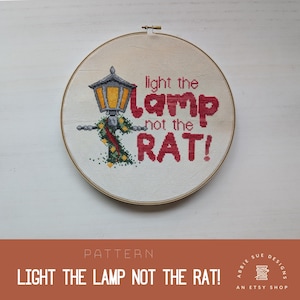 May include: A cross-stitch pattern featuring a lamppost with a red and green garland and the text "light the lamp not the rat!"