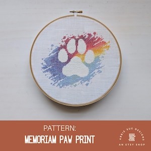 Może przedstawiać: Wzór haftu krzyżykowego z białą odbitką łapy na tle akwarelowym w kolorach tęczy. Wzór nosi tytuł "MEMORIAM PAW PRINT" i jest dostępny w sklepie Abbie Sue Designs na Etsy.