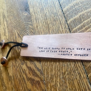 May include: A copper bookmark with a quote by Audrey Hepburn engraved on it. The quote reads, "The best thing to hold onto in life is each other." The bookmark has a black leather strap with two copper rivets.