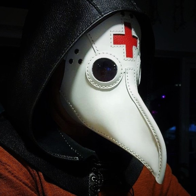 May include: A white plague doctor mask with a long beak and a red cross on the forehead. The mask has a black leather hood attached to it.