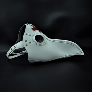 May include: White leather plague doctor mask with a red cross on the beak and a round eye hole.