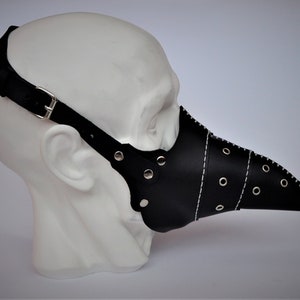 May include: A black leather plague doctor mask with a long, beak-like nose. The mask has white stitching, silver rivets, and adjustable black straps with a silver buckle. The mask is displayed on a white mannequin head.