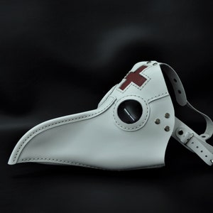 May include: White leather plague doctor mask with a red cross on the forehead and a black eye piece. The mask has a long beak and is secured with a leather strap.