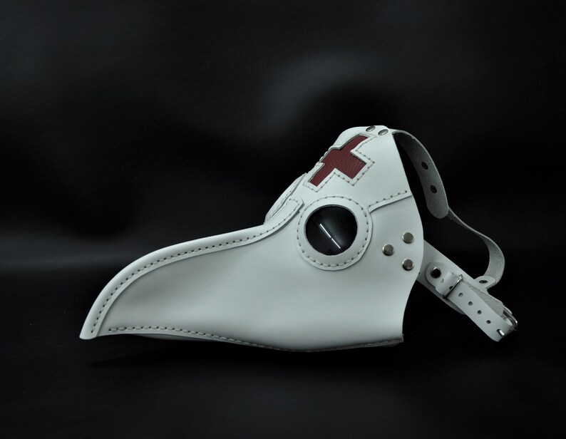 May include: White leather plague doctor mask with a red cross on the forehead and a black eye piece. The mask has a long beak and is secured with a leather strap.