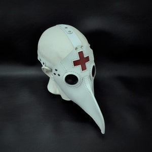 May include: White leather plague doctor mask with a red cross on the beak. The mask has two round eye holes and a long, pointed beak.