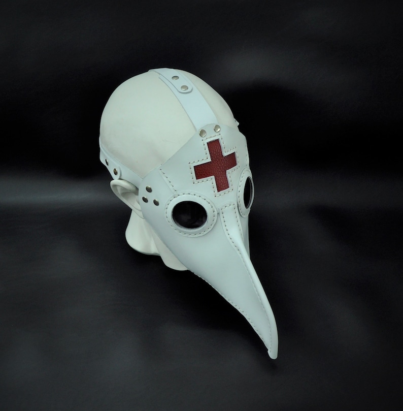May include: White leather plague doctor mask with a red cross on the beak. The mask has two round eye holes and a long, pointed beak.