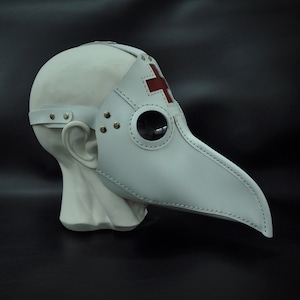 May include: White leather plague doctor mask with a red cross on the forehead and a round, clear eye piece. The mask has a long, pointed beak.