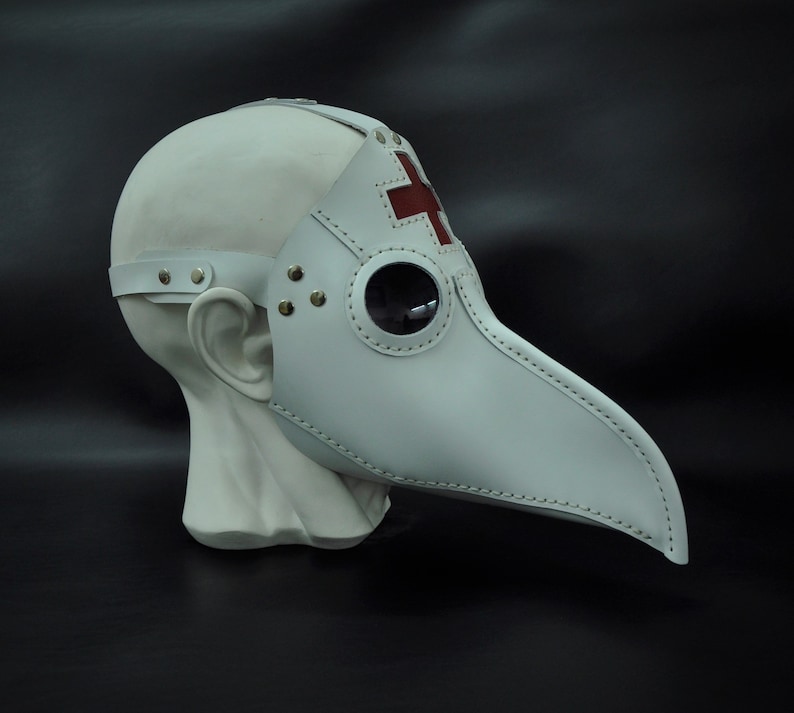 May include: White leather plague doctor mask with a red cross on the forehead and a round, clear eye piece. The mask has a long, pointed beak.