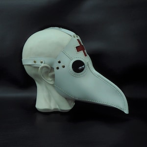 May include: White leather plague doctor mask with a red cross on the forehead and a stitched beak. The mask is on a white mannequin head.