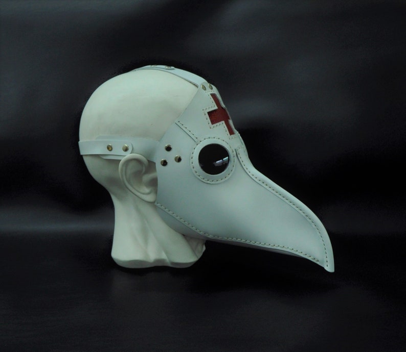 May include: White leather plague doctor mask with a red cross on the forehead and a stitched beak. The mask is on a white mannequin head.