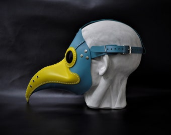 May include: A blue and yellow leather plague doctor mask with a long beak and eye holes. The mask is worn on a mannequin head.