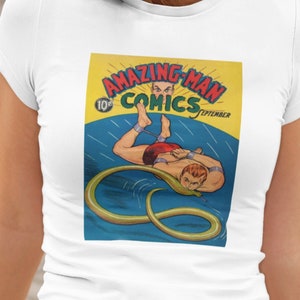 Op de afbeelding: Een wit T-shirt met een vintage comicboekomslag bedrukt op de voorkant. De omslag toont een man in een rode zwembroek die wordt aangevallen door een slang. De comicboekomslag is getiteld "Amazing-Man Comics" en kost 10 cent. De comicboekomslag is gedateerd september.