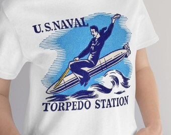 Op de afbeelding: Wit T-shirt met een blauwe en witte afbeelding van een matroos die op een torpedo rijdt met de tekst "U.S. NAVAL TORPEDO STATION".