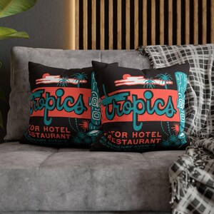 Peut inclure: Deux coussins noirs avec un design r&eacute;tro "Tropics" en turquoise et corail. Les coussins comportent le texte "Motor Hotel Restaurant" et "418 McHenry Avenue". Les coussins sont sur un canap&eacute; gris.