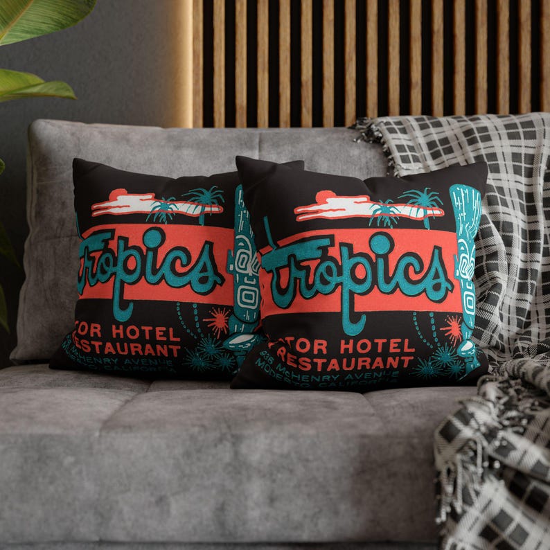 Peut inclure: Deux coussins noirs avec un design r&eacute;tro "Tropics" en turquoise et corail. Les coussins comportent le texte "Motor Hotel Restaurant" et "418 McHenry Avenue". Les coussins sont sur un canap&eacute; gris.