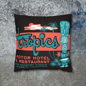 Peut inclure: Coussin carr&eacute; noir avec un design r&eacute;tro. Il pr&eacute;sente le mot "Tropics" en script turquoise sur un rectangle corail. Comprend une figure tiki et le texte d'un motel et restaurant &agrave; Modesto, Californie.