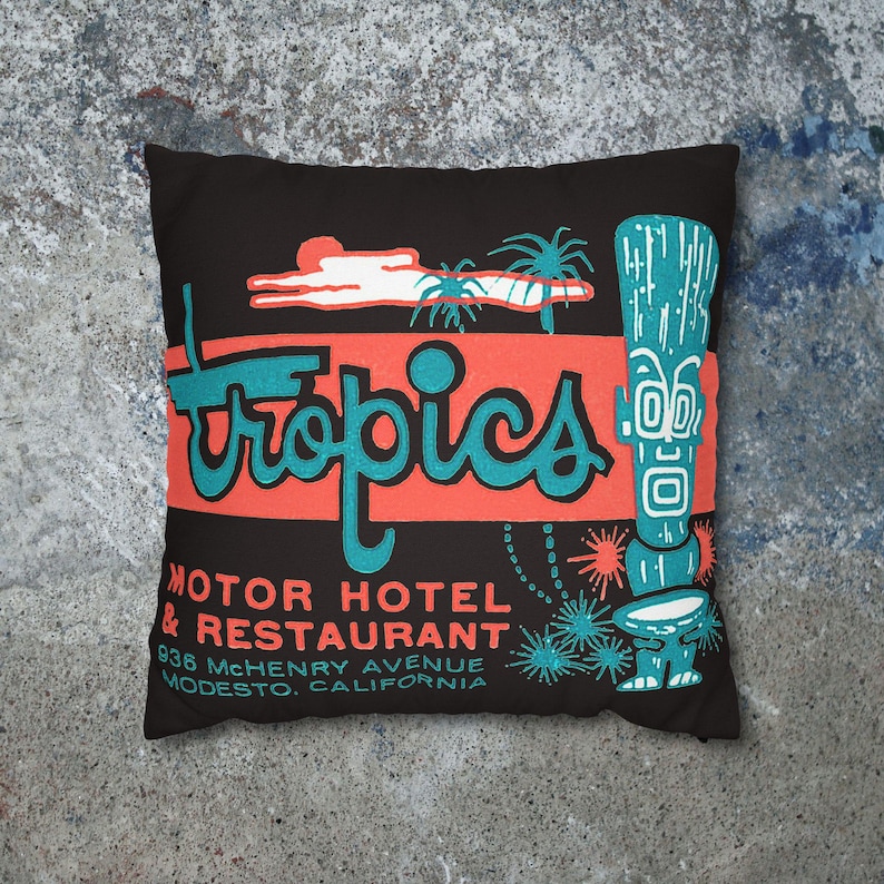 Peut inclure: Coussin carr&eacute; noir avec un design r&eacute;tro. Il pr&eacute;sente le mot "Tropics" en script turquoise sur un rectangle corail. Comprend une figure tiki et le texte d'un motel et restaurant &agrave; Modesto, Californie.