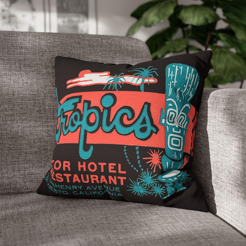 Peut inclure: Coussin noir avec un design r&eacute;tro avec le mot "Tropics" en script turquoise sur un rectangle rouge. Le coussin comprend &eacute;galement un tiki turquoise et des palmiers, avec du texte pour un restaurant d'h&ocirc;tel.